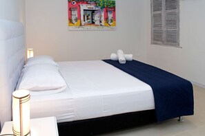 A neatly made bed with white linens and a dark blue blanket, a bedside lamp, and a wall painting of a colorful building. - Beautiful 3 Bedroom Old City House (Cartagena de Indias)