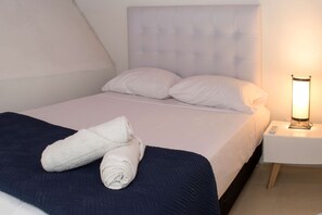 Un lit bien fait, avec une tête de lit blanche, une couvre-lit bleue et deux serviettes blanches roulées posées sur le lit. On aperçoit une table de chevet sur laquelle se trouve une lampe.