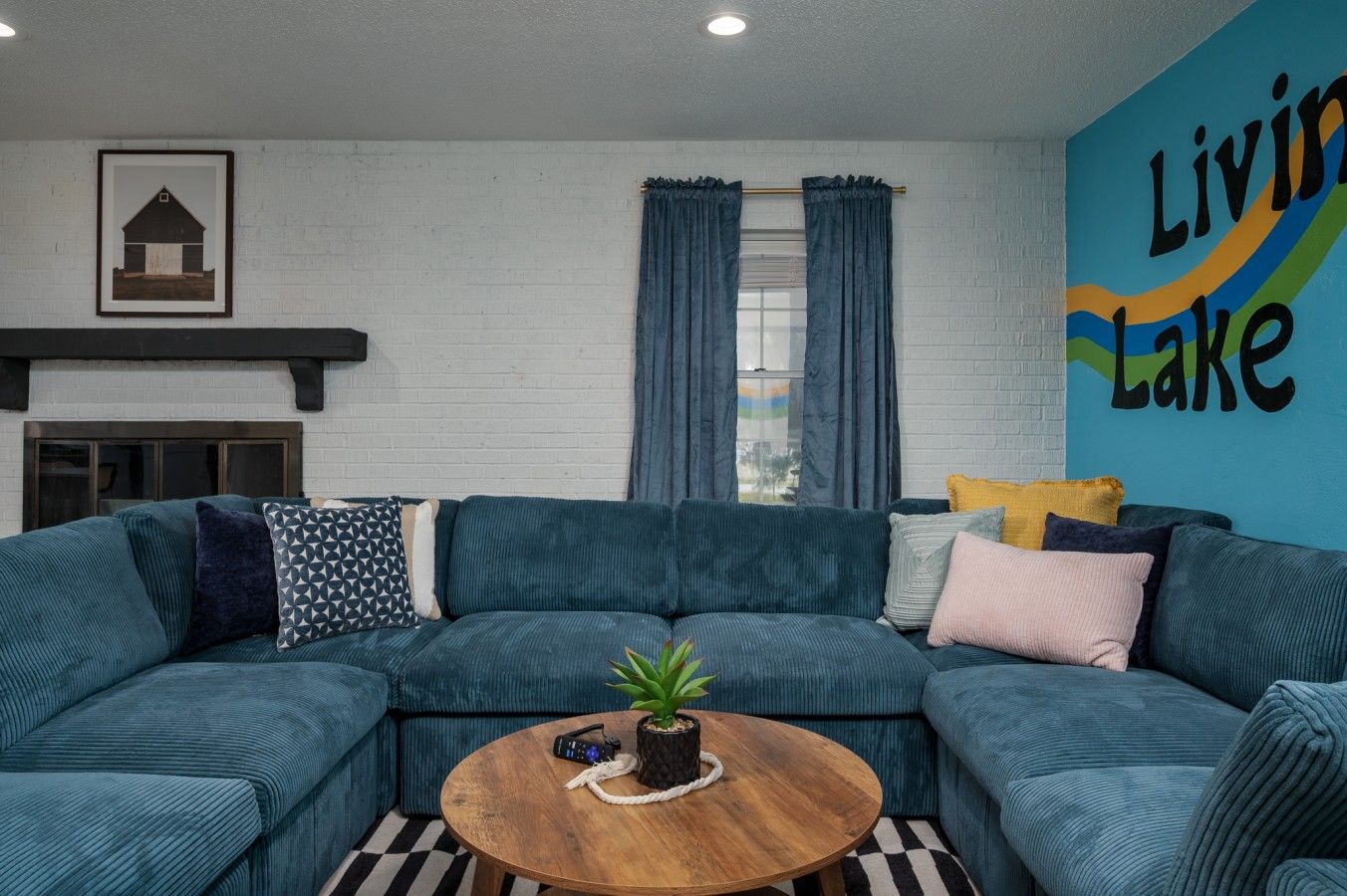 A living room with a sectional sofa, a coffee table with a plant, a fireplace, and a wall with the text 'Living Lake'.