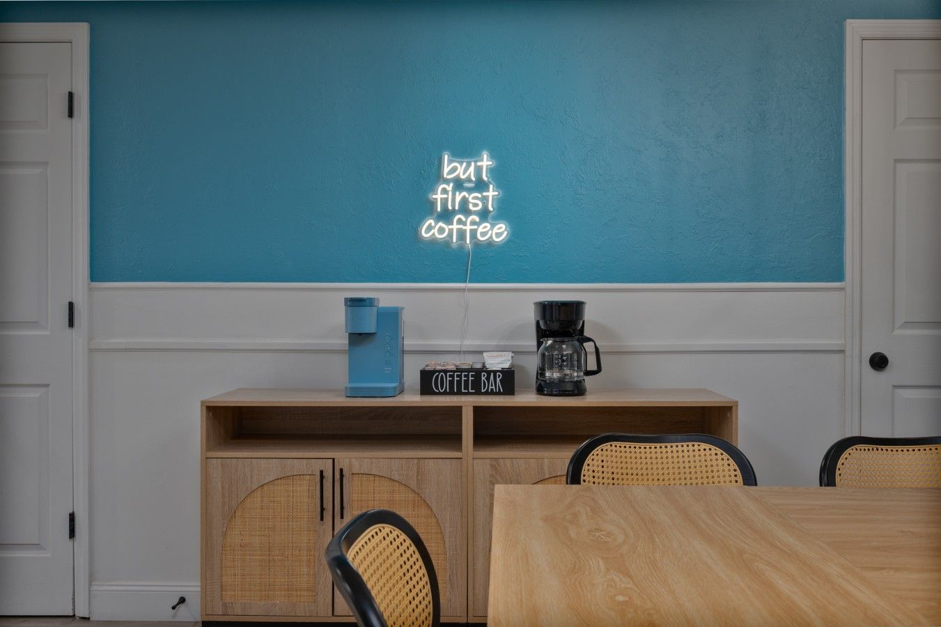 A room with a teal wall, a coffee bar with a sign, a blue coffee maker, and a black coffee maker.