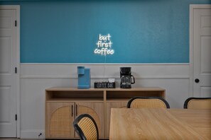 A room with a teal wall, a coffee bar with a sign, a blue coffee maker, and a black coffee maker.