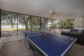 An outdoor ping pong table with a ceiling fan, a brick wall, and a view of trees and water.