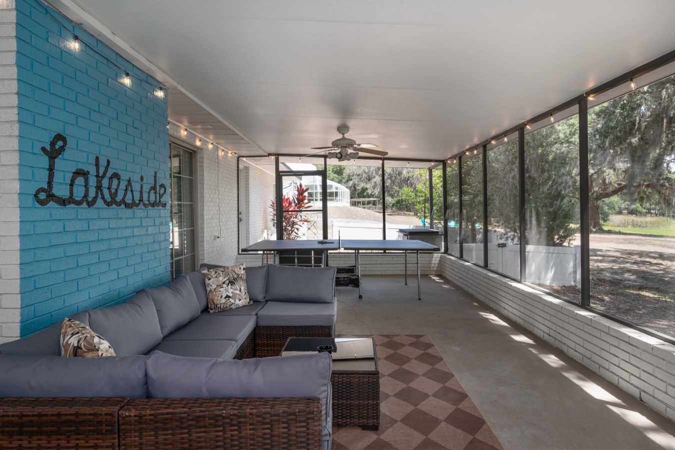 A covered patio area with a blue brick wall, a gray sectional sofa, a table, and chairs.