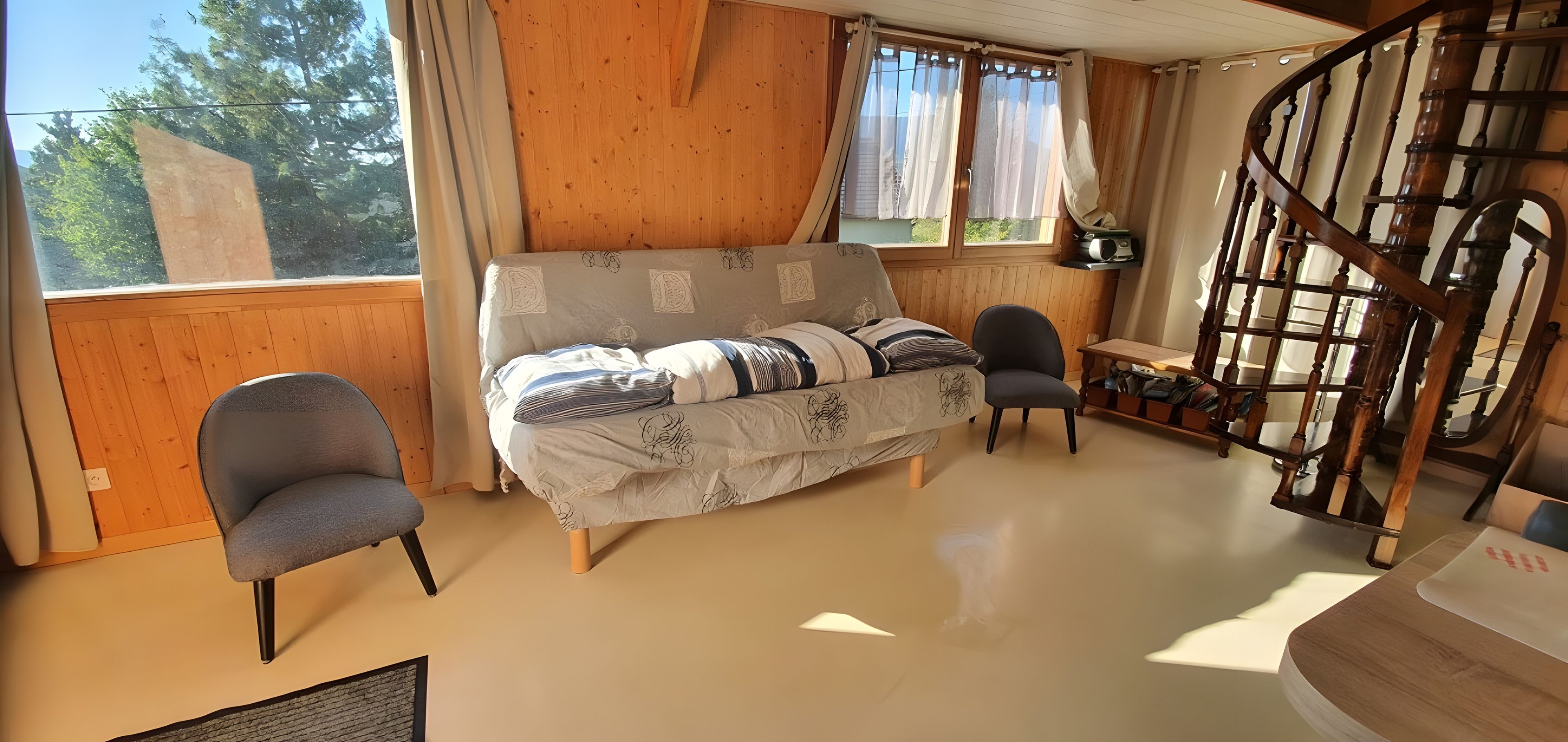 Ein Zimmer mit Holzvertäfelung, einer Couch mit gemusterten Kissen, zwei Stühlen, einer Holztreppe und einem Fenster mit Vorhängen.