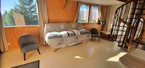 Een kamer met houten inrichting, een bank met gestreepte kussens, twee stoelen, een houten trap en een raam met gordijnen.