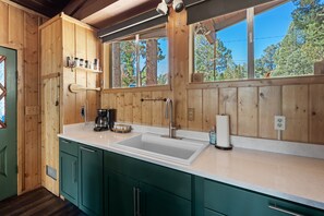 Une cuisine rustique, située dans une cabane en bois, avec une grande fenêtre, un évier et des armoires vertes.