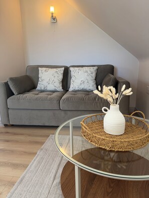 Un salon moderne avec un canapé gris, des coussins à motifs floraux, une table basse en verre surmontée d’un panier en osier et un vase contenant des plantes séchées.