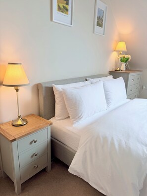 A neatly made bed with white bedding, a bedside lamp, a nightstand with a drawer, and a wall-mounted lamp with a vase of flowers. - 1 Bedroom Private Meadow View Annexe Apartment (Allington)