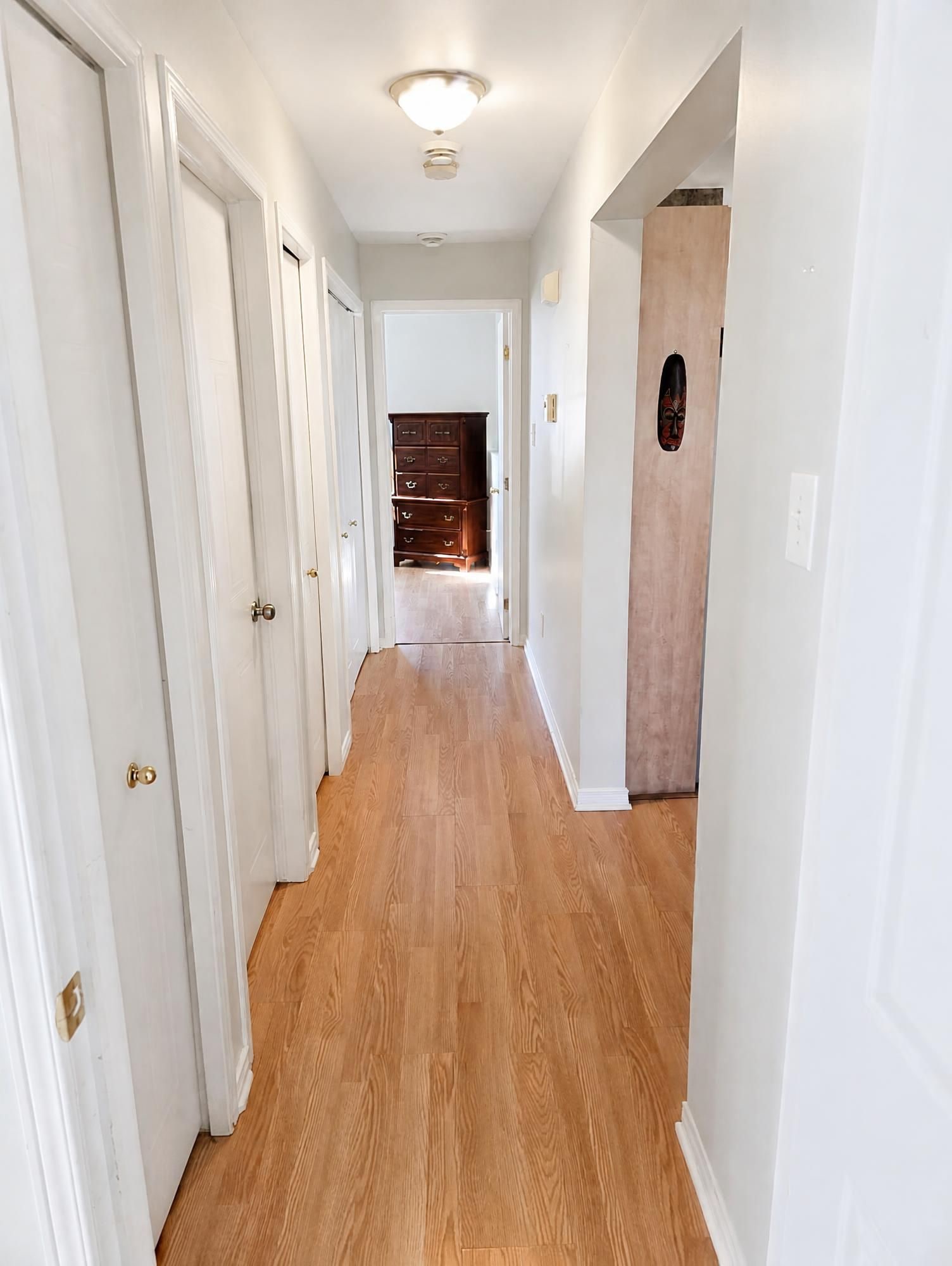 Un pasillo con suelo de madera, paredes blancas y una puerta al final que da a otra habitación.