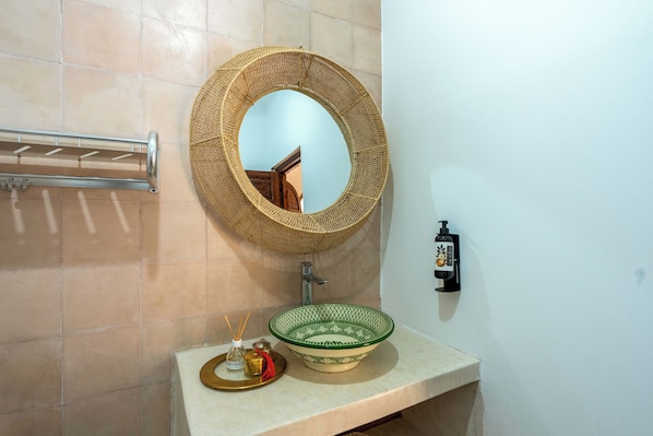A bathroom with a round woven mirror, a green patterned bowl sink, a towel rack, and a soap dispenser. - Dar Nouraï (marrakech)