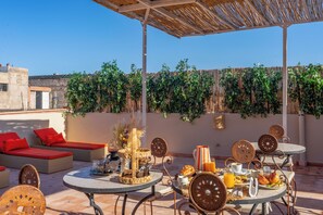 Terrassa al terrat amb seients a l'aire lliure, taula de dinar preparada per l'esmorzar, i un fons de vegetació i cel blau.
