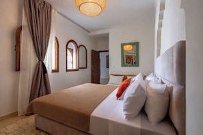 A hotel room with a bed, curtains, a wooden door, a mirror, and a wall-mounted picture. - Dar Nouraï (marrakech)