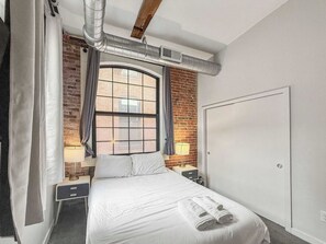 Een hotelkamer met een groot bed, een raam met gordijnen, een bakstenen muur en een witte kledingkast.