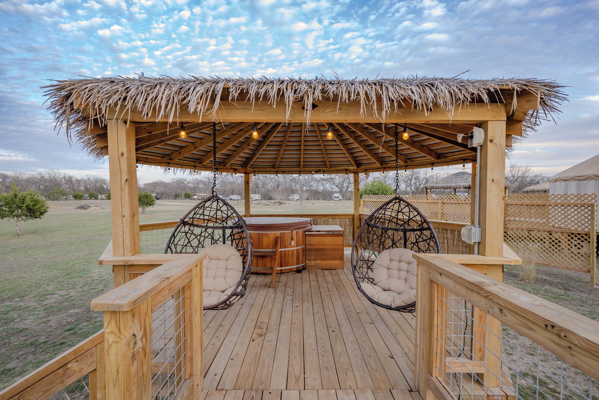A wooden deck with a thatched roof, two hanging chairs, and a wooden table.