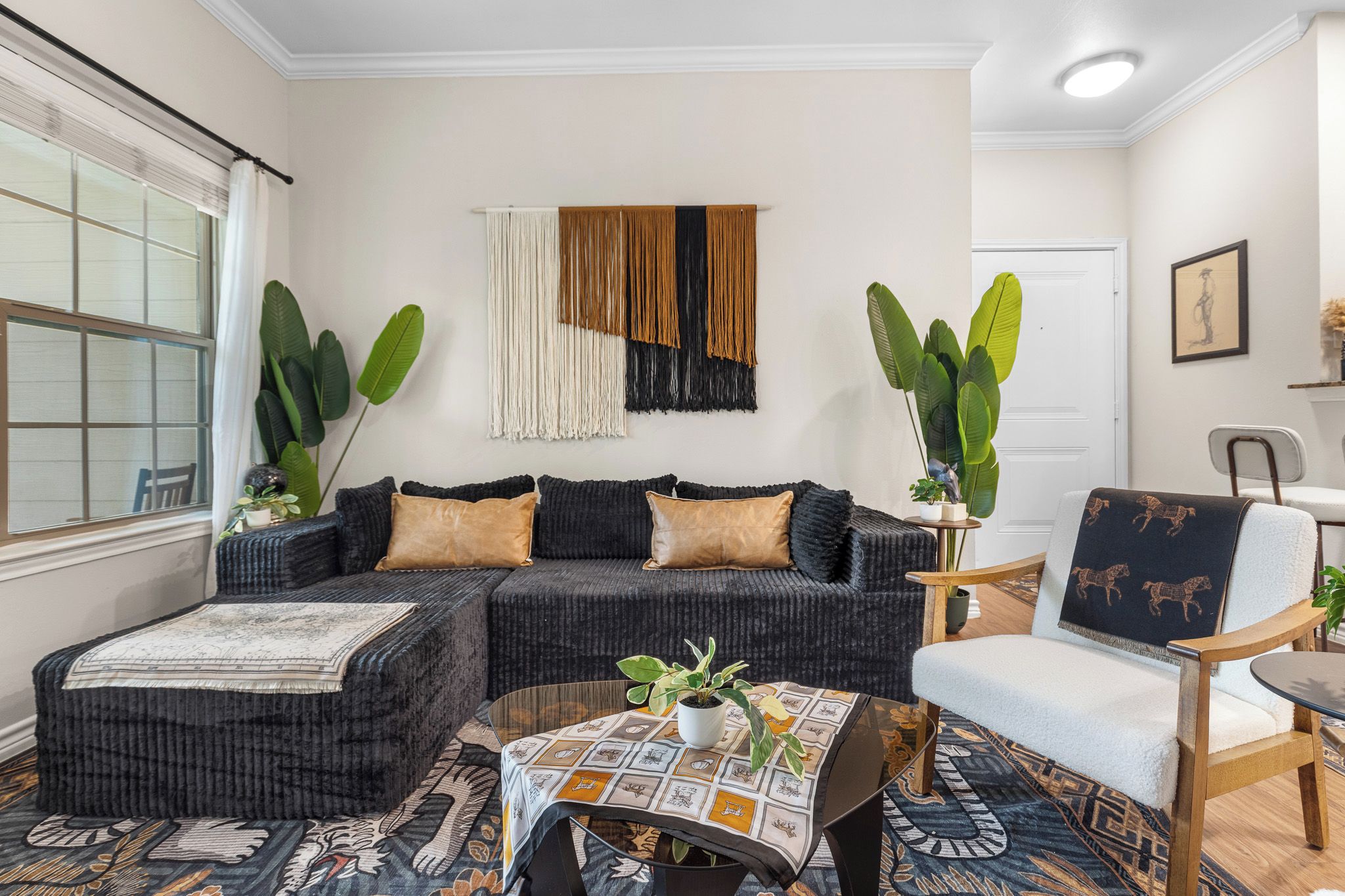 Ein Wohnzimmer mit einem Ecksofa, einem runden Couchtisch und einem Sessel. Es gibt ein großes Fenster mit Vorhängen, eine Wanddekoration aus Makramee und einen Blumentopf.