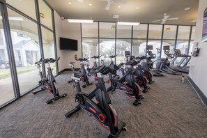 Ein Fitnessraum mit in Reih und Glied stehenden Ergometer, großen Fenstern und einem Deckenventilator.