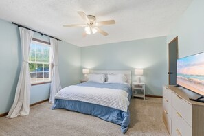 Ein Schlafzimmer mit einem Bett, zwei Nachttischen, einem Deckenventilator, einem Fenster mit Vorhängen und einem Fernseher auf einer Kommode.