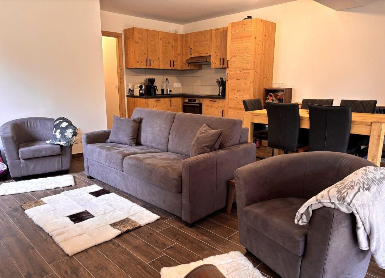 A living room with a grey sofa, a smaller armchair, a coffee table, and a rug. There is a wooden cabinet with a stove and a dining table with chairs.
