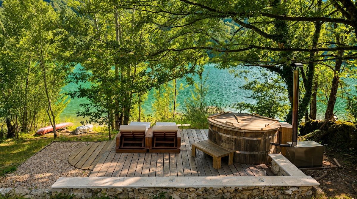 Eine Holzterrasse mit Sitzgelegenheit im Freien und einem Whirlpool, umgeben von Bäumen und Blick auf einen See.