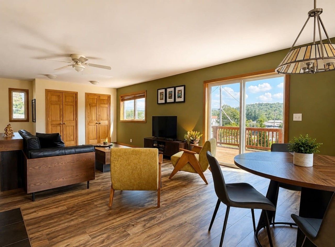 A modern living room with a sofa, chairs, a dining table, a TV, and a balcony with a view.
