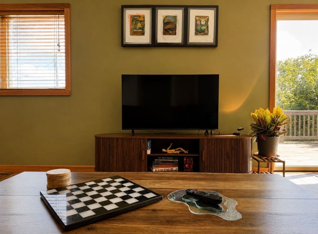 A room with a large flat-screen TV mounted on the wall, a wooden table with a chessboard, a remote control, and a potted plant.