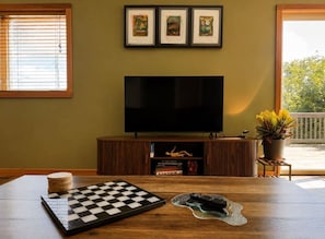 A room with a large flat-screen TV mounted on the wall, a wooden table with a chessboard, a remote control, and a potted plant. - 5th Street Trail House- 3Bed- 2Bath (Davis)