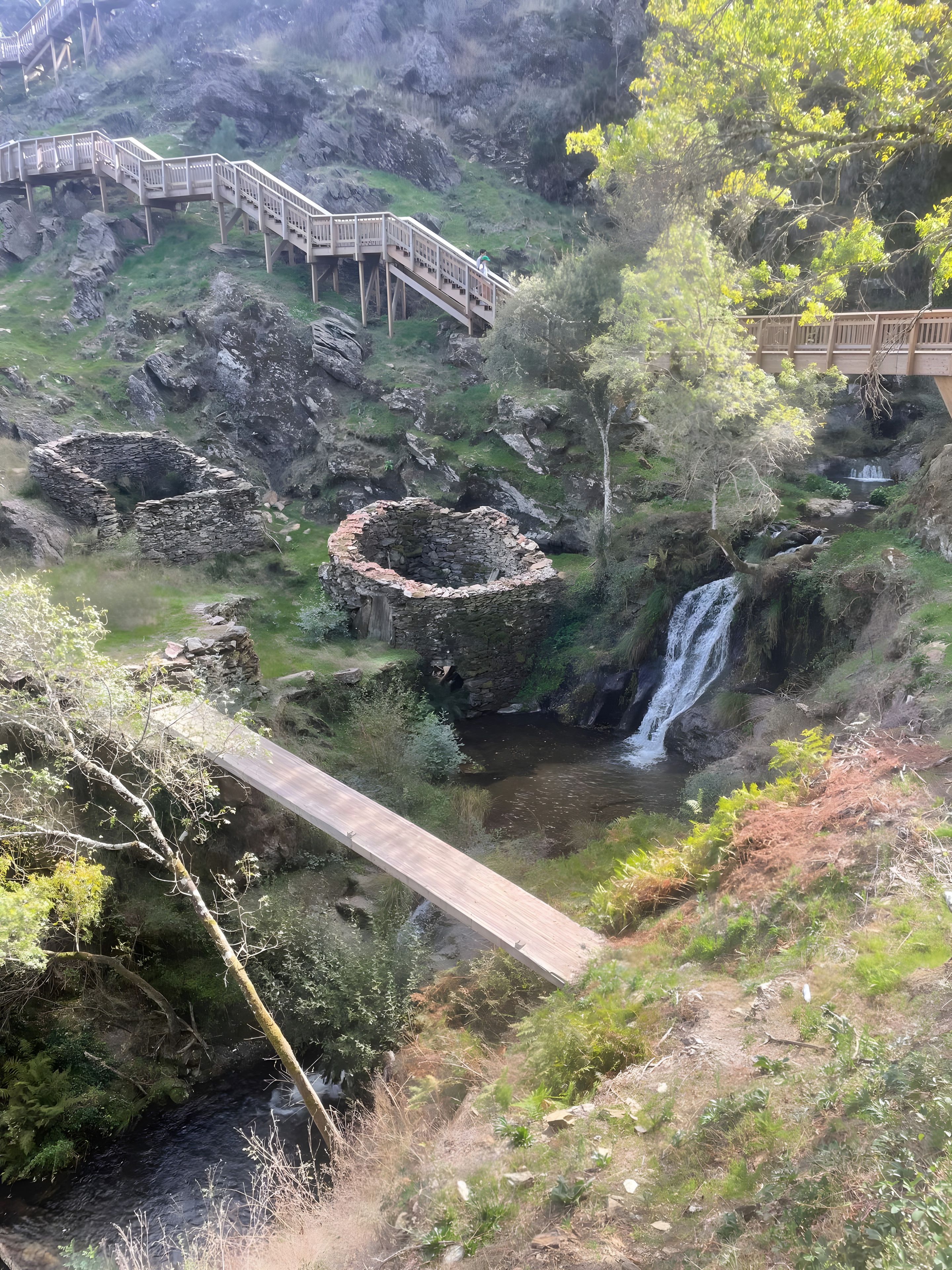 Eine Steinstruktur mit einer Holzbrücke, einem Wasserfall und einem bewaldeten Gebiet.