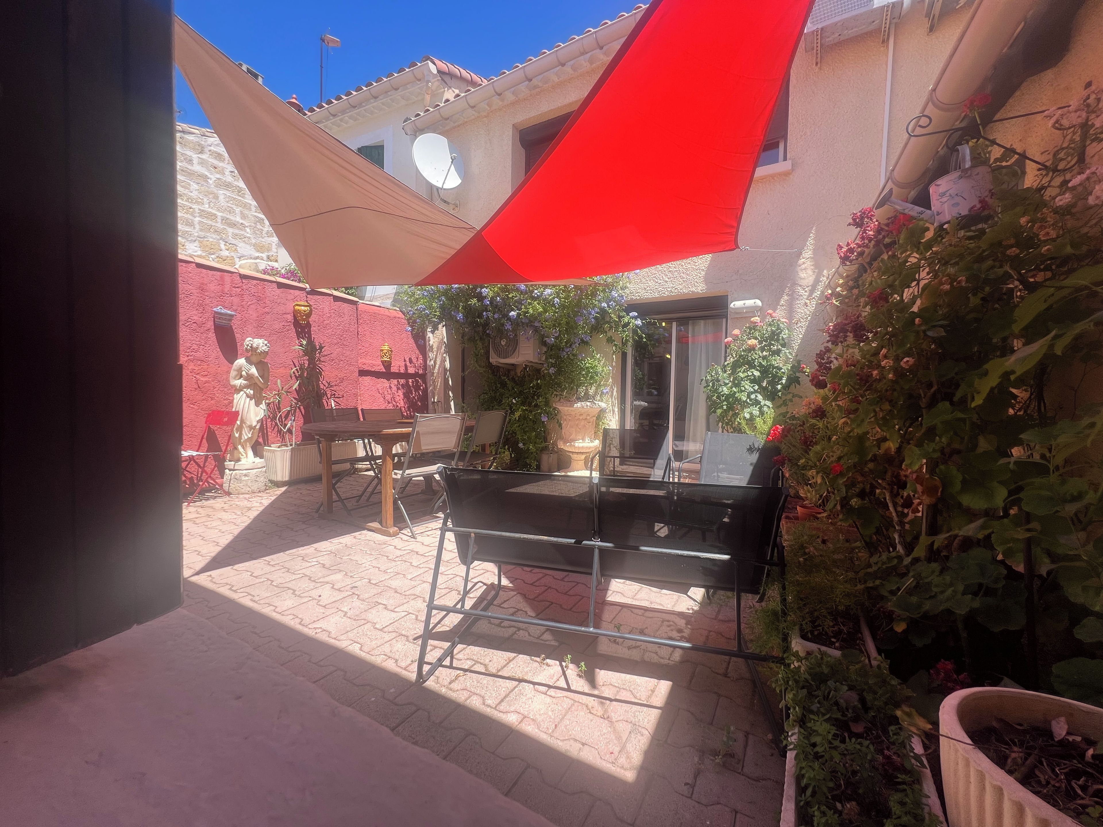 Een binnentuin met een rood en beige zonnescherm, tuinmeubels, potplanten en een standbeeld.