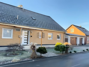En bostadsbyggnad med en fasad av tegel, ett tak täckt med takpannor och en liten trädgård framför huset med olika buskar och ett träd.