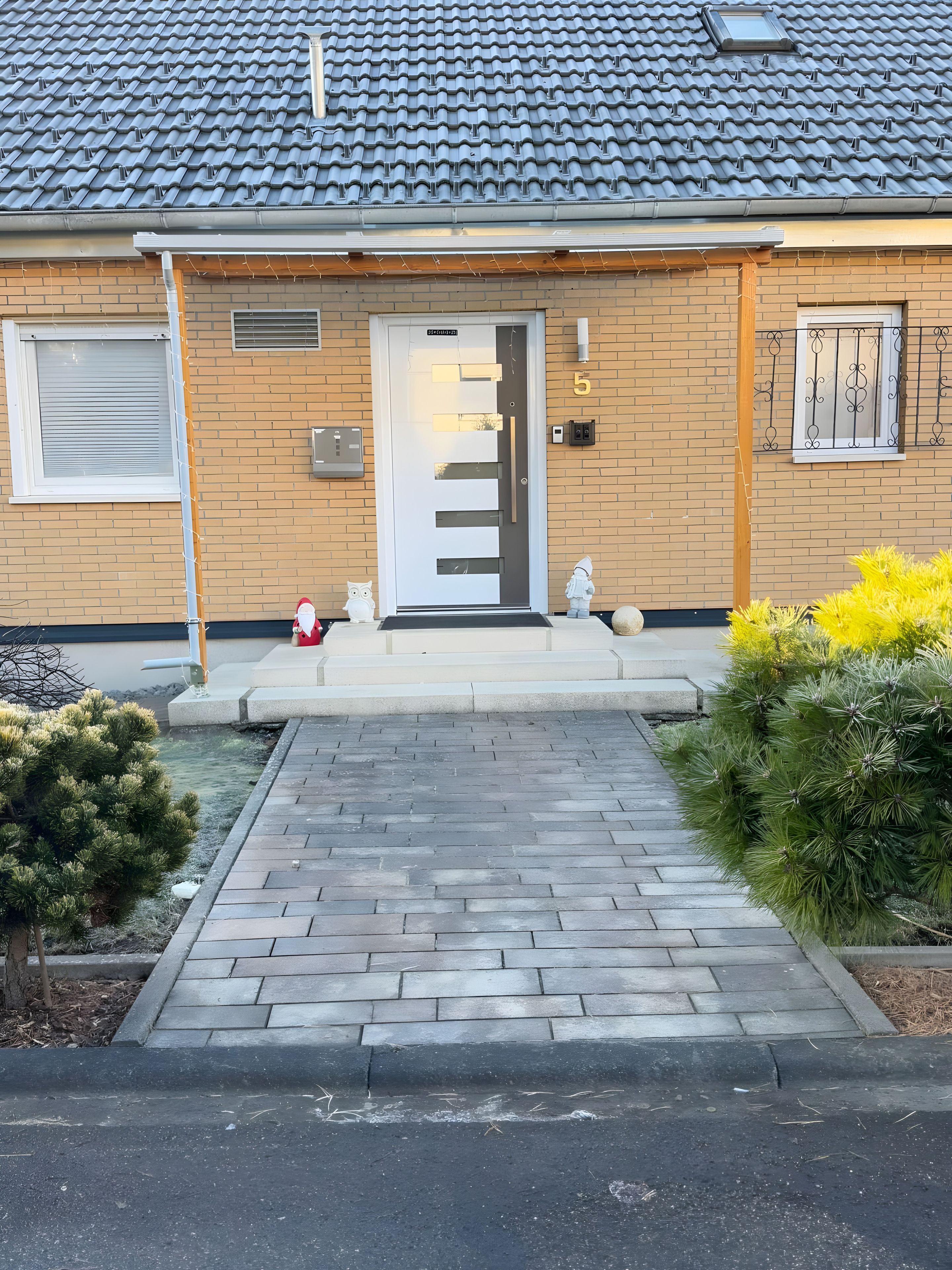 En bostadsbyggnad med ett tak täckt av takpannor, en ytterdörr och en liten trädgårdsyta med buskar.