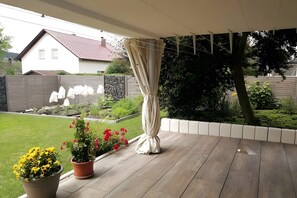 Uma área de varanda coberta com piso de madeira, plantas em vasos e uma cortina branca.