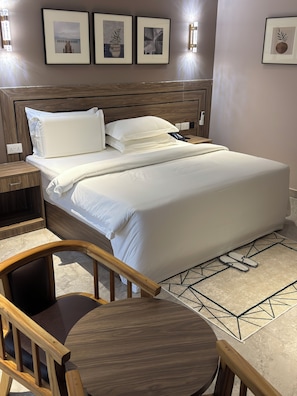 A neatly made bed with white linens, a wooden headboard, and a bedside table with a lamp. There are framed pictures on the wall above the bed.