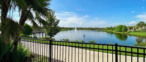 A serene lake with a fountain, surrounded by lush greenery and a clear blue sky.