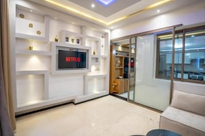 Un saló modern amb televisió de pantalla plana penjada a una paret blanca, una porta de vidre que dona a la cuina i un sofà beix.