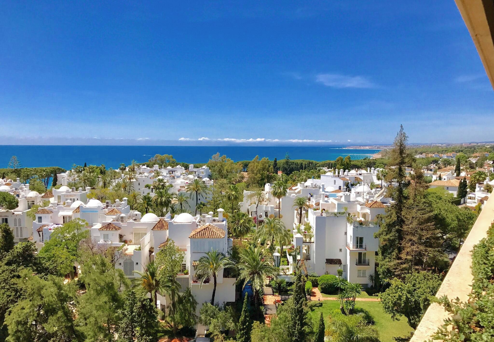 A coastal residential area with white buildings, palm trees, and a clear view of the sea.
