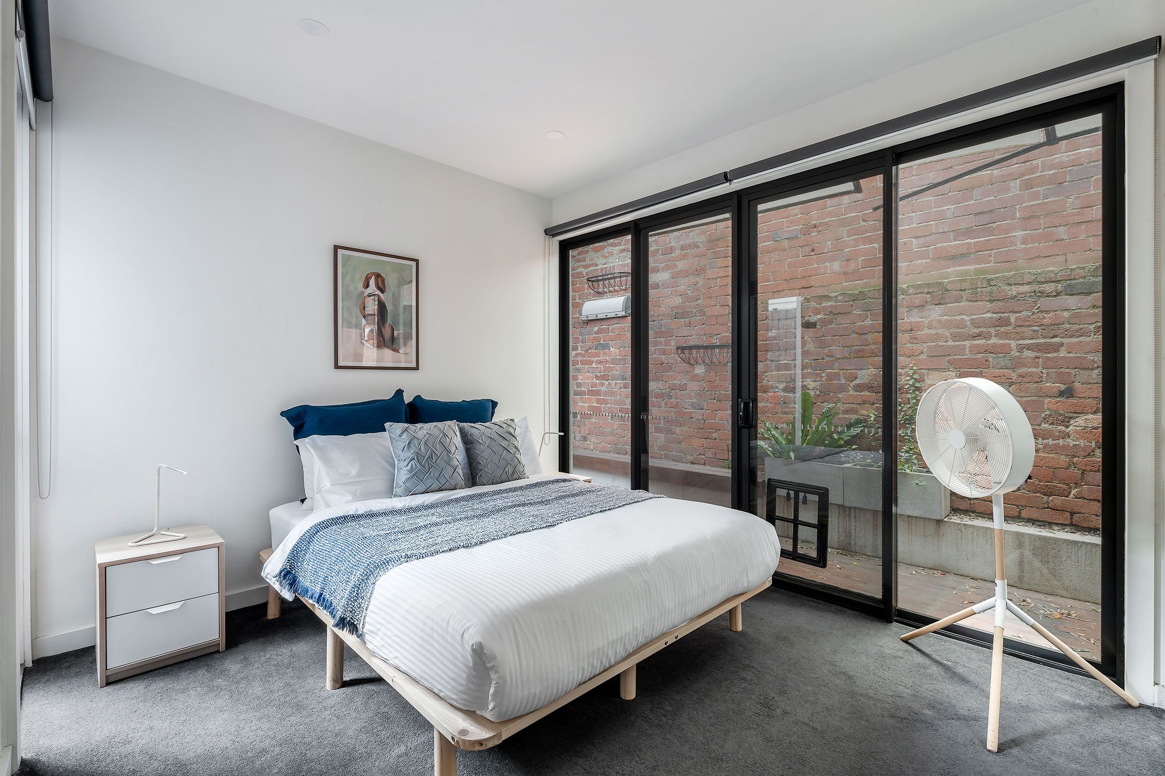A bedroom with a bed, a nightstand, a radiator, a window with a view of a brick wall, and a fan.