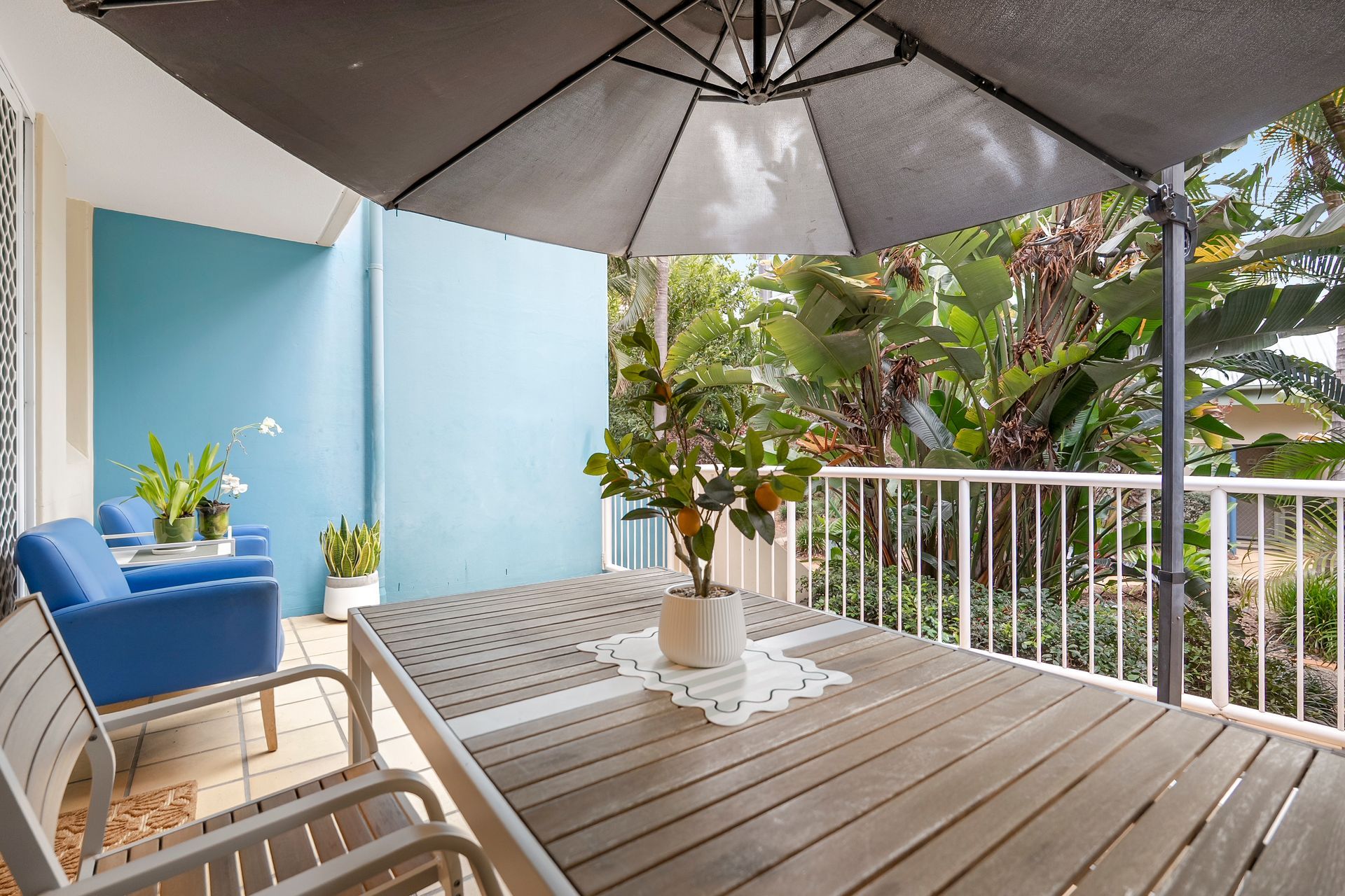An outdoor patio with a wooden table, blue chairs, a white umbrella, and potted plants.