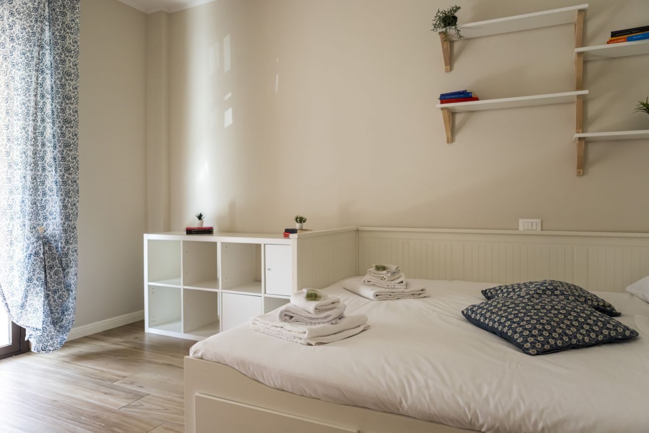 Een slaapkamer met een bed, nachtkastje, kast en een plank met boeken en een plant.