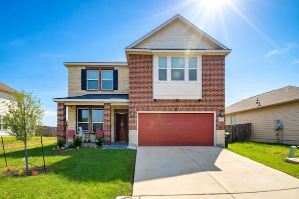 A two-story residential house with a red garage door, a brick facade, and a small front yard with a tree and a patio table. - Game Night HQ! Arcade, Pool Table & 5BR (Seguin)