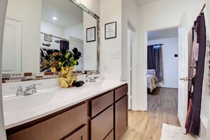 A bathroom with a large mirror, a vanity with a sink, and a decorative vase with flowers. - Game Night HQ! Arcade, Pool Table & 5BR (Seguin)