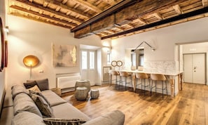 A spacious living area with a wooden ceiling, a sofa set, a dining table with bar stools, and a large window. - DESIGNER ROMAN APARTMENT WITH TWO ENSUITE BEDROOMS AND A SPA-LIKE TOUCH (Roma)