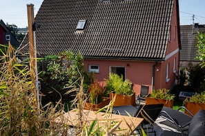 A red house with a tiled roof, a small window, and a door. There are plants in front of the house and a chair to the right. - New Listing (Schwarzenbach am Wald)