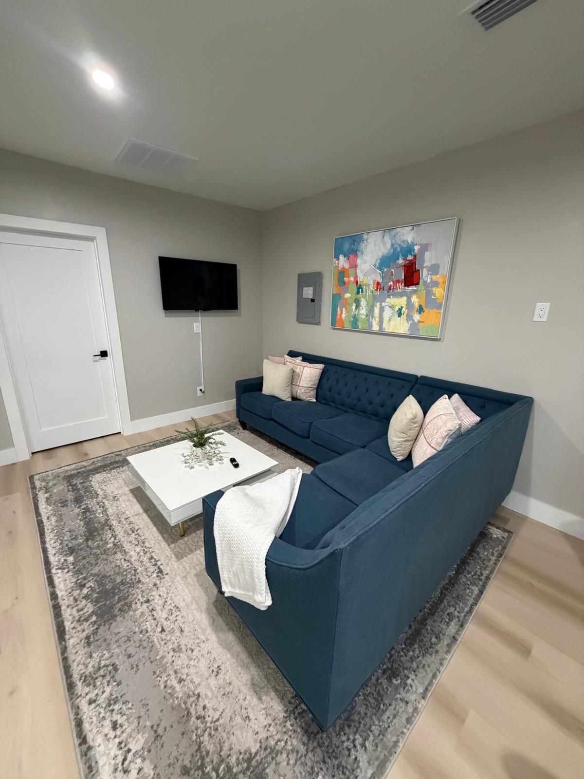 A living room with a blue sofa, a white coffee table, a gray rug, a flat-screen TV mounted on the wall, and a painting above the sofa.