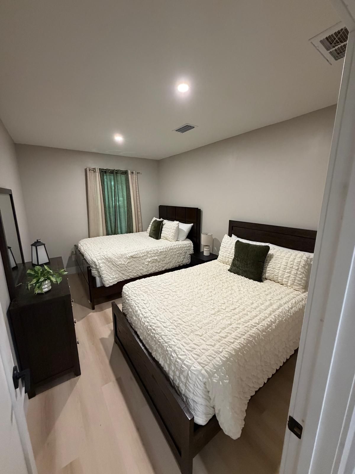 A modern bedroom with two beds, a bedside table, a mirror, and a window with curtains.