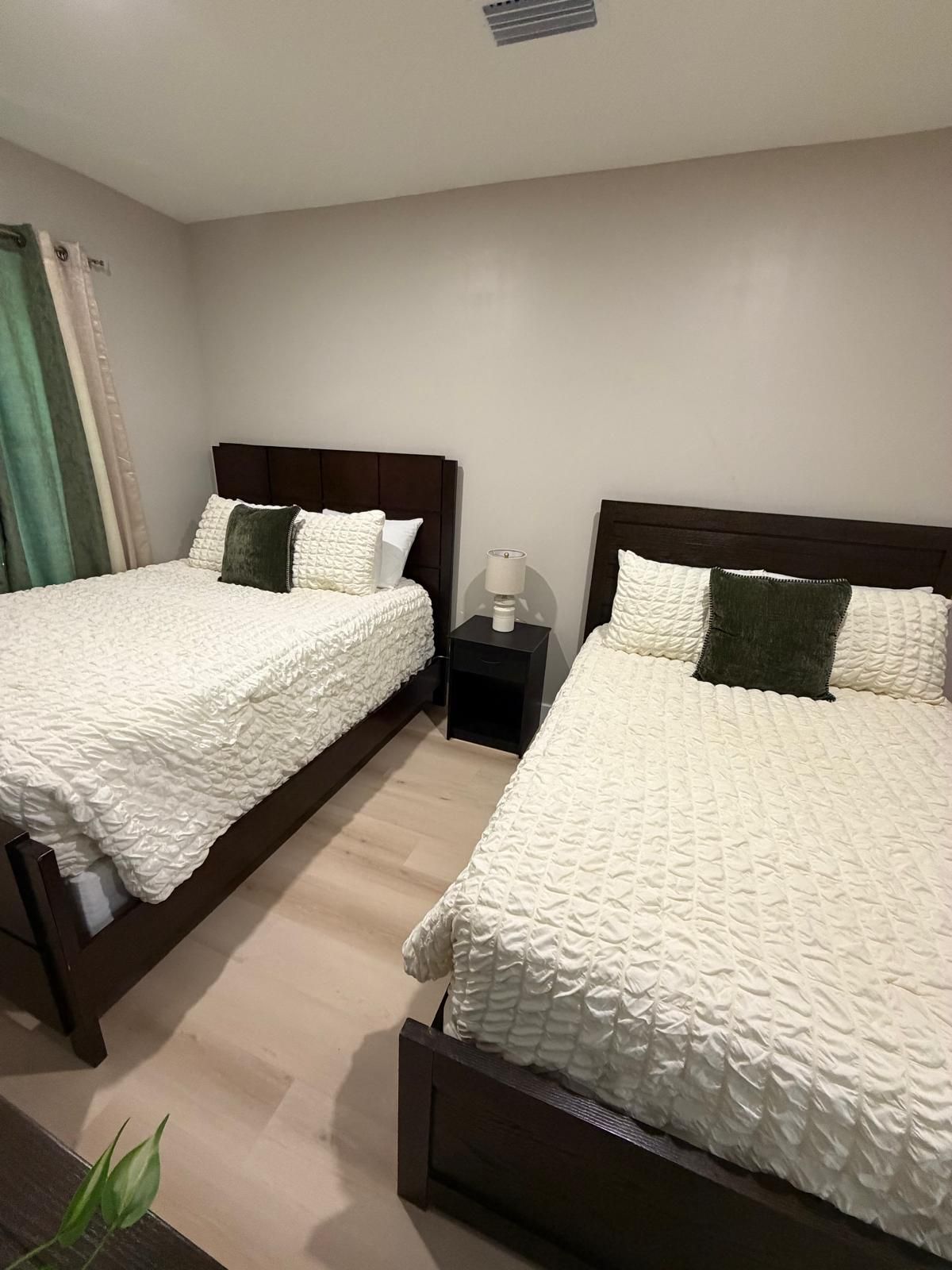 A hotel room with two beds, each with a dark headboard and white textured bedding. There is a nightstand with a lamp between the beds.
