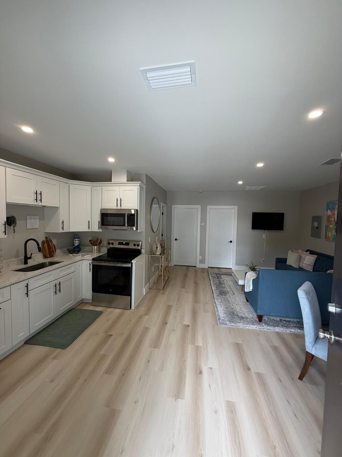 A modern kitchen with white cabinets, a microwave, and a stove. There is a dining area with a blue sofa and a TV mounted on the wall.