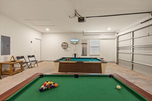 Una mesa de billar en una sala con proyector en el techo, una televisión y una ventana con persianas.