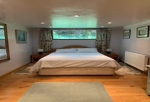 A bedroom with a large bed, wooden flooring, a window with curtains, and framed pictures on the walls. - Secret, private log cabin in large beautiful  gardens, very quiet, pet allowed (Norwich)