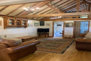 A spacious living room with a high wooden ceiling, a television, leather sofas, a patterned rug, and a wooden cabinet. - Secret, private log cabin in large beautiful  gardens, very quiet, pet allowed (Norwich)
