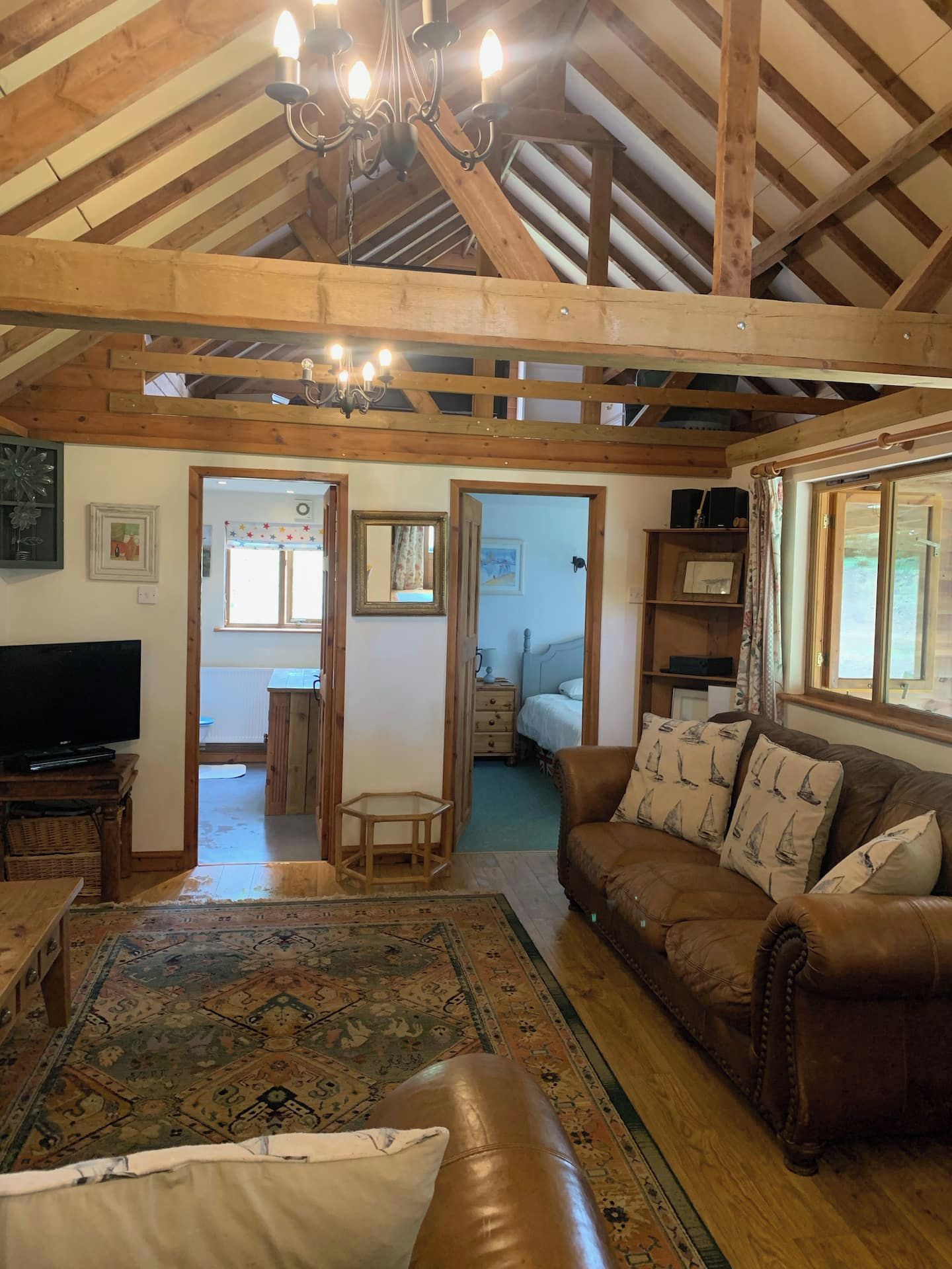 A spacious living room with a high ceiling, wooden beams, a chandelier, a leather sofa, a television, and a patterned rug.